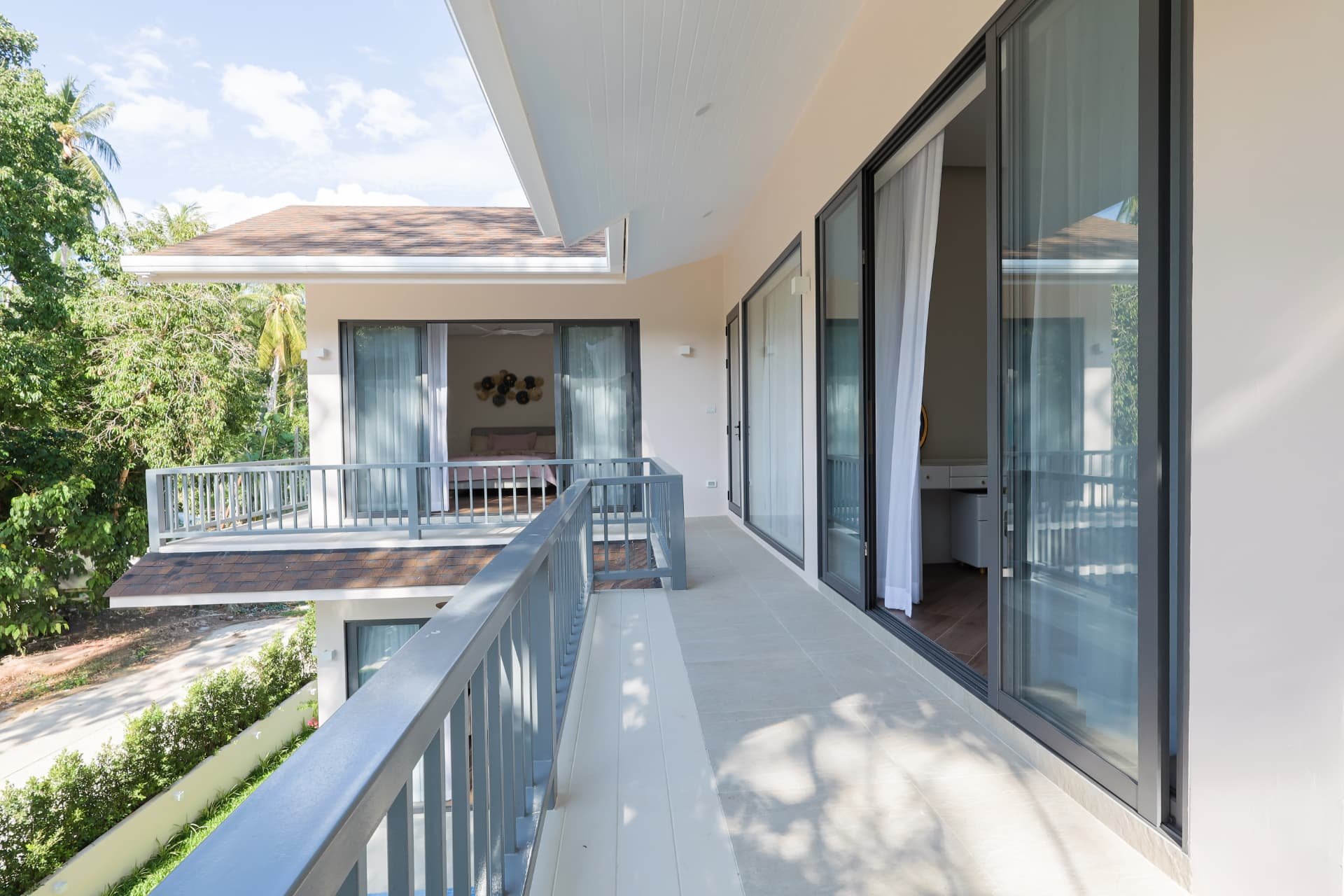 Upper-floor balcony running along the villa with sliding glass doors to the bedrooms, a grey railing and views over the tropical garden