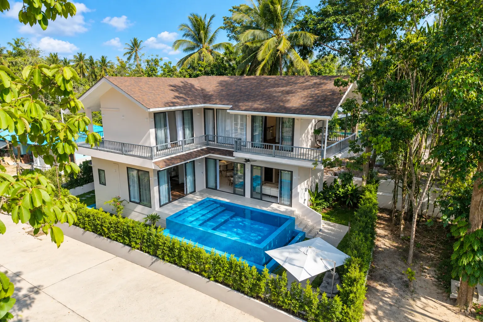 Daytime aerial view of the villa with its glass-sided swimming pool, hedged garden and coconut palms