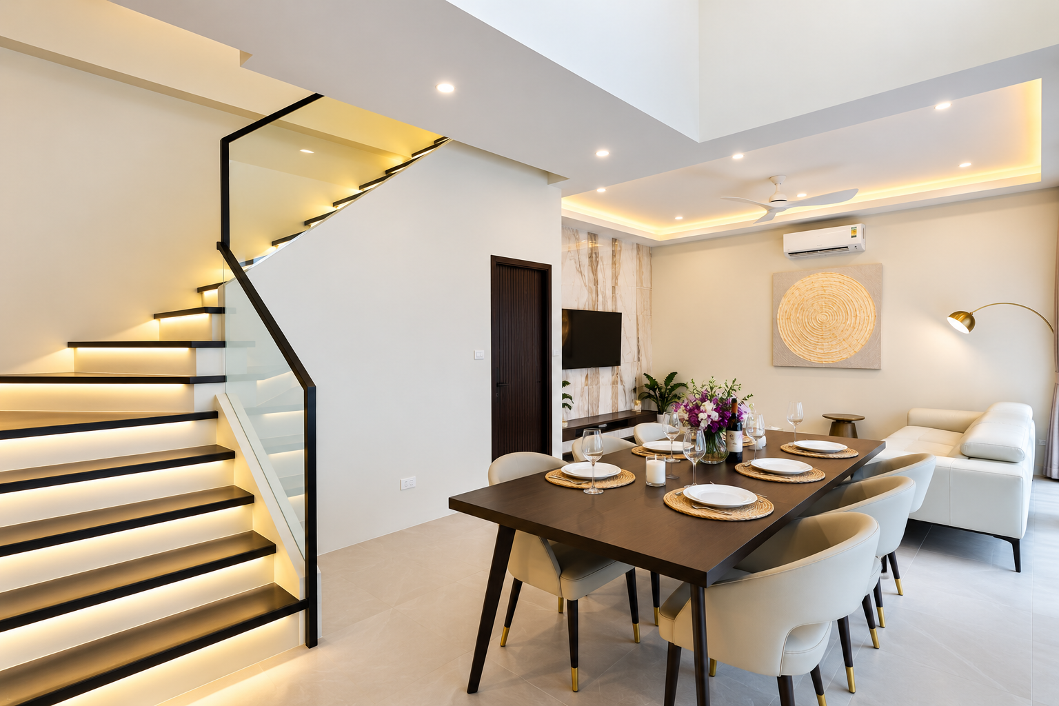 Dining table set for eight next to the illuminated staircase, with the living area and sofa behind