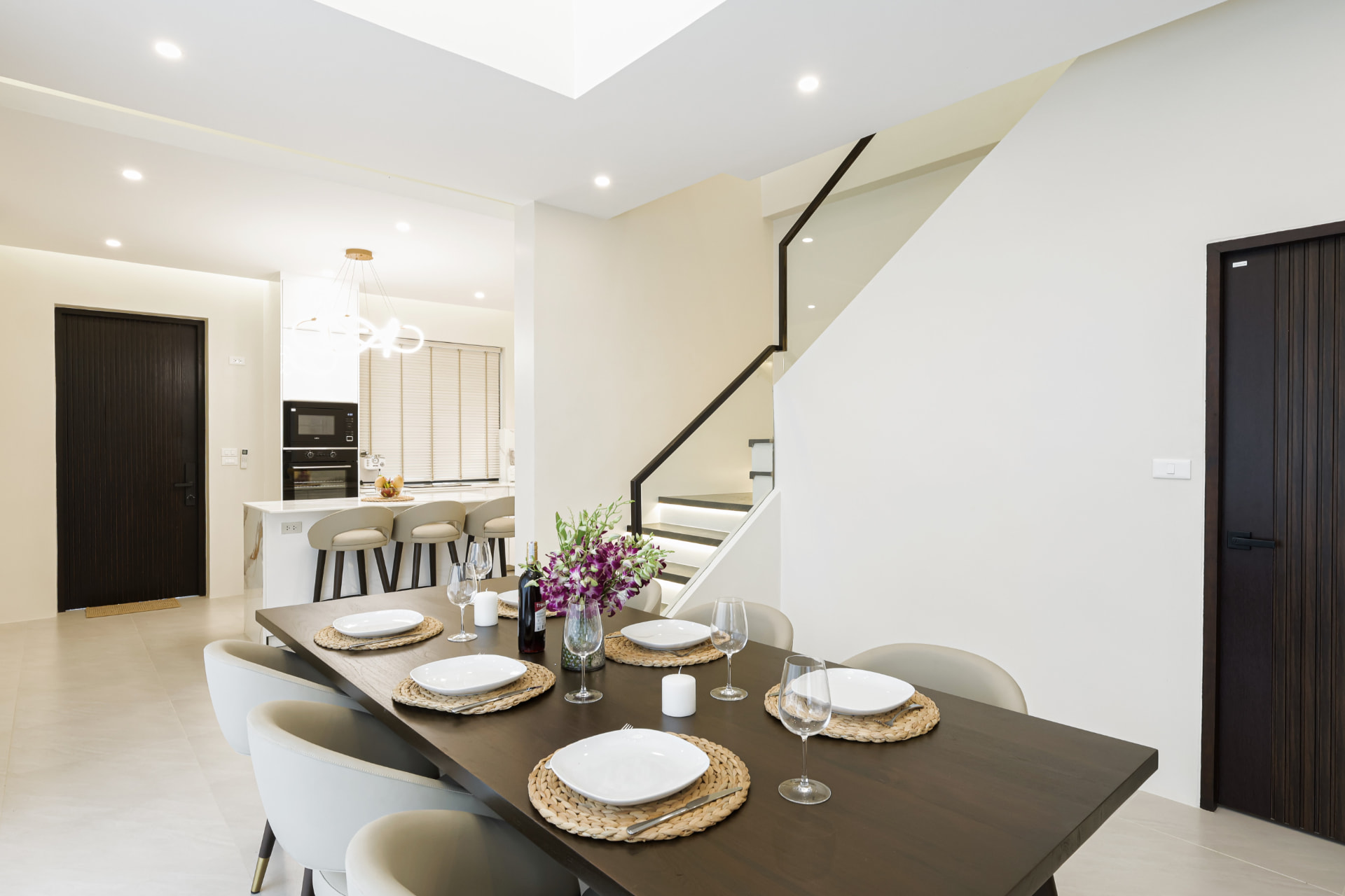 Dining table set for six beside the staircase, with the open kitchen and breakfast bar behind